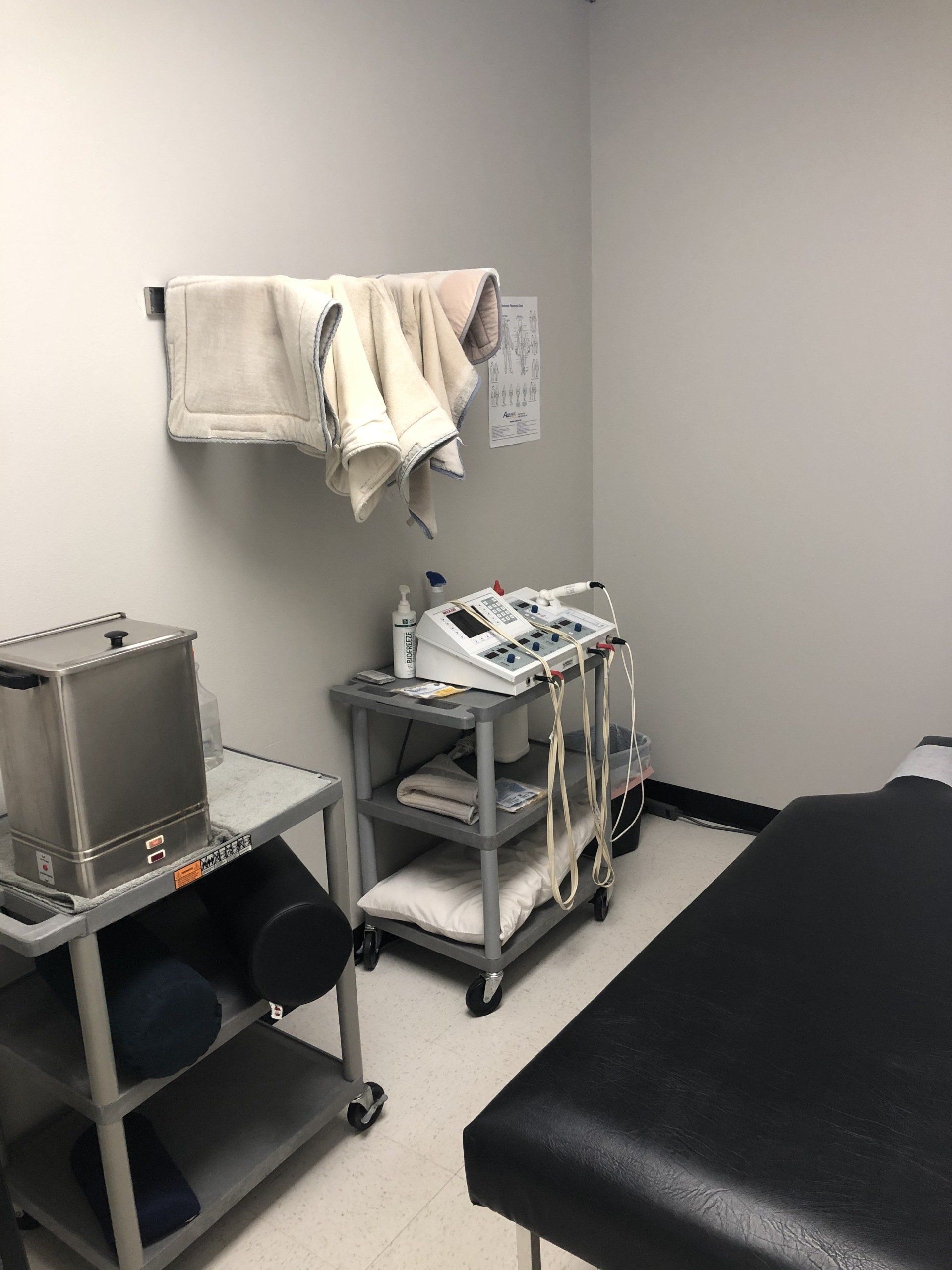 Physical therapy room with equipment: tables, cart with a machine, towels on wall, and treatment bed.