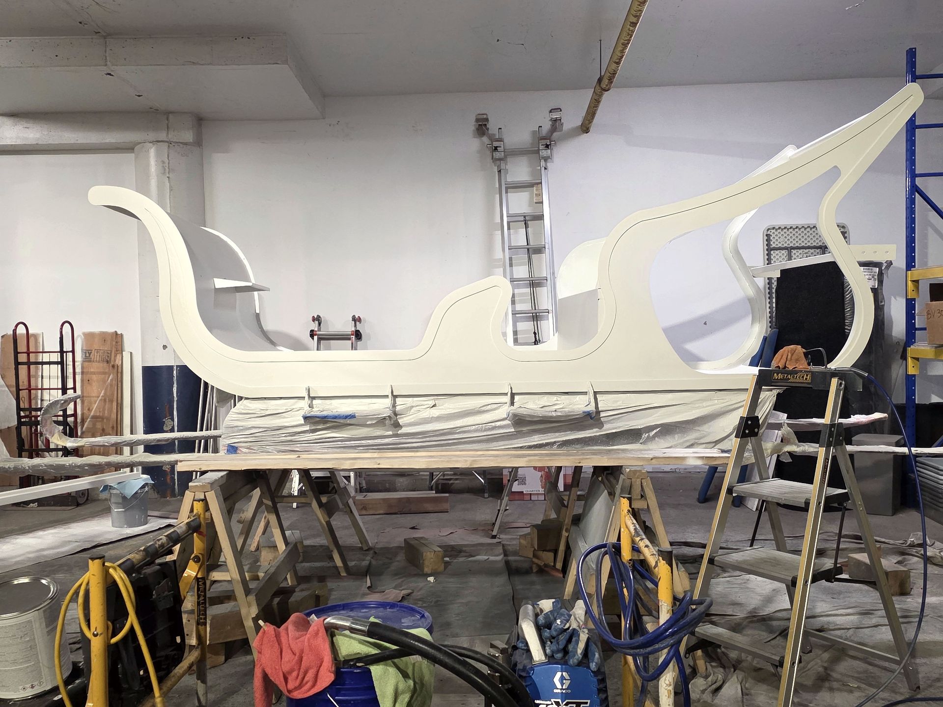 Coque de bateau blanche en construction dans un atelier, entourée d'échelles, d'outils et d'échafaudages.