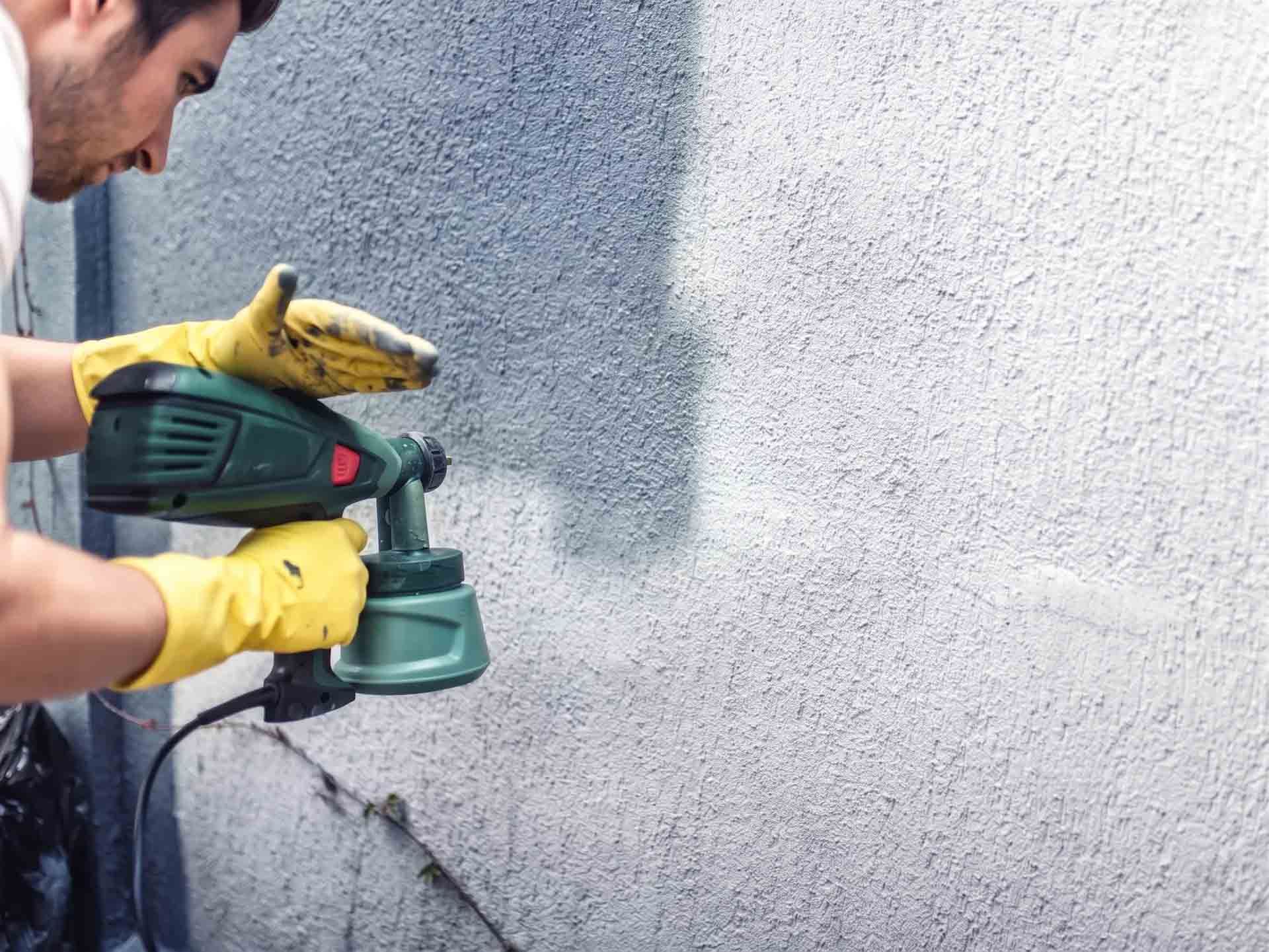 Personne utilisant une ponceuse électrique verte sur un mur gris, portant des gants jaunes à l'extérieur