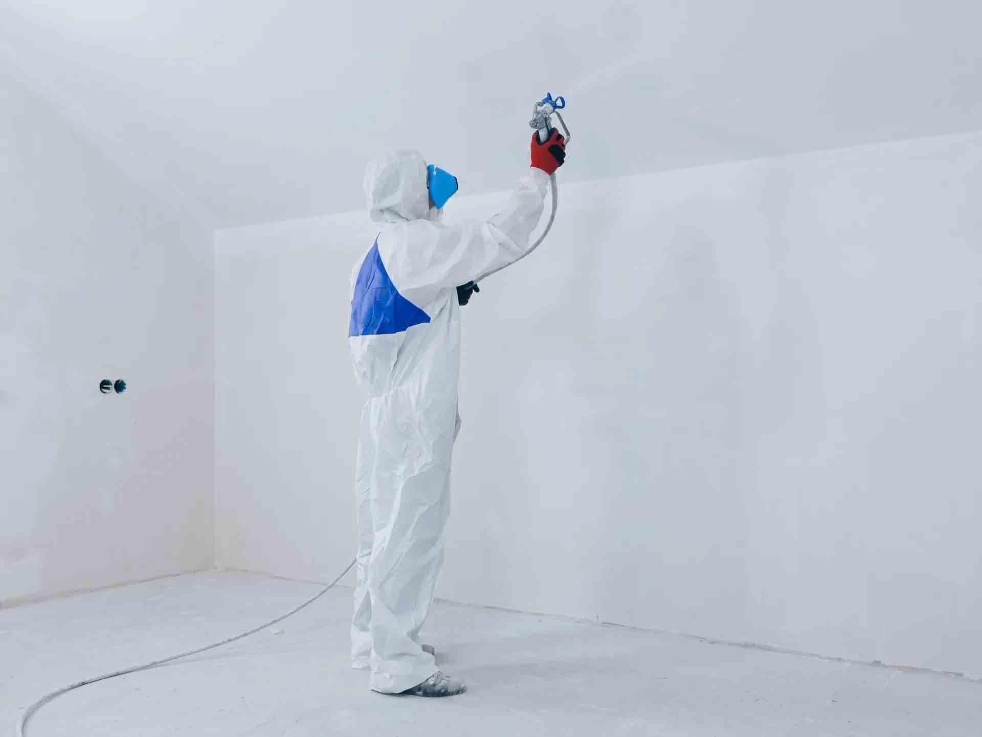 Un ouvrier en combinaison de protection blanche peint au pistolet le plafond d'une pièce blanche.