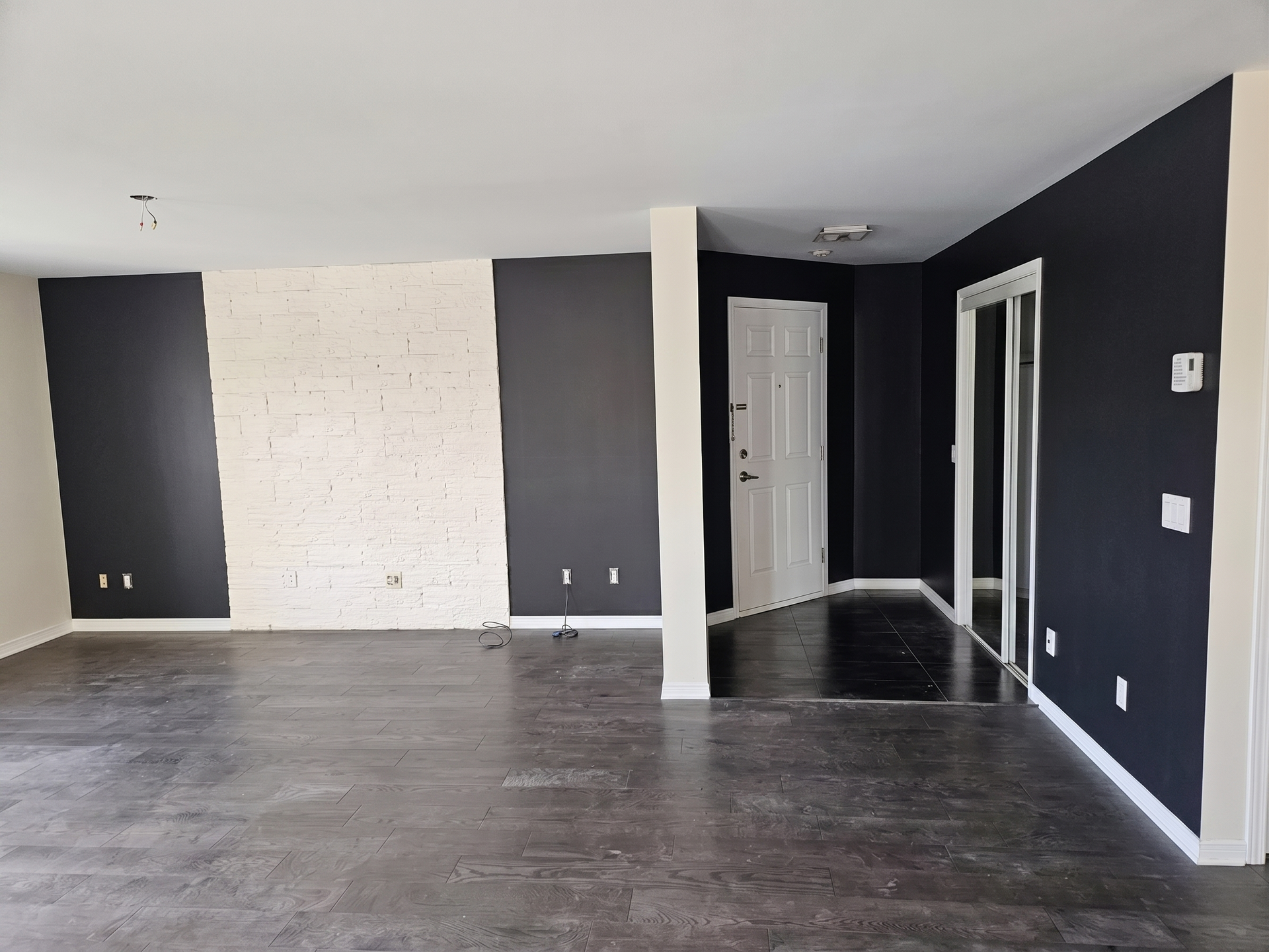 Salon moderne vide avec des murs d'accent sombres, une cheminée en briques blanches et un parquet gris.
