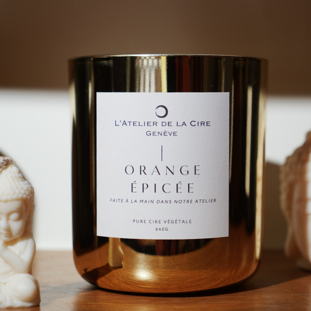 Bougie dorée dans un pot avec étiquette blanche « Orange Épicée ». Placée près de statues de Bouddha blanches.