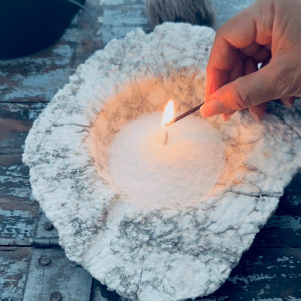 Allumage manuel d'une bougie dans un bol blanc à la texture rugueuse. La flamme vacillante illumine la bougie.