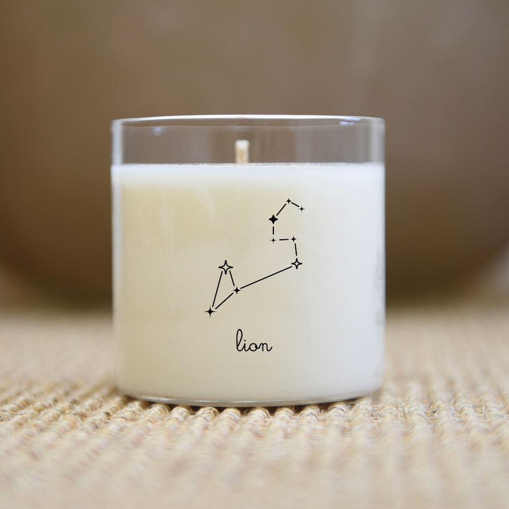 Bougie blanche dans un pot en verre avec une illustration de la constellation du Lion noire sur une surface texturée.