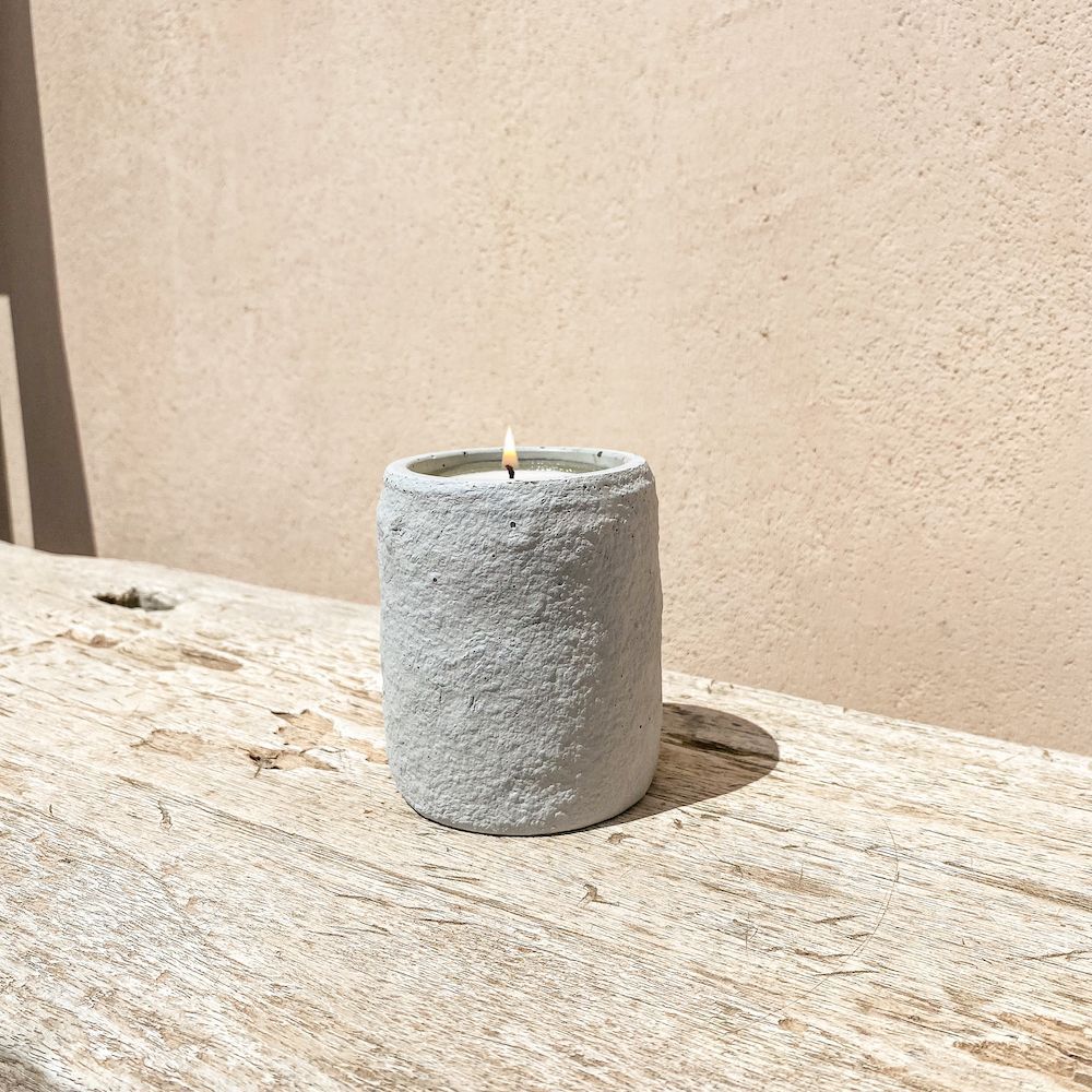 Bougie allumée dans un support en béton gris texturé sur une surface en bois avec un mur beige en arrière-plan.