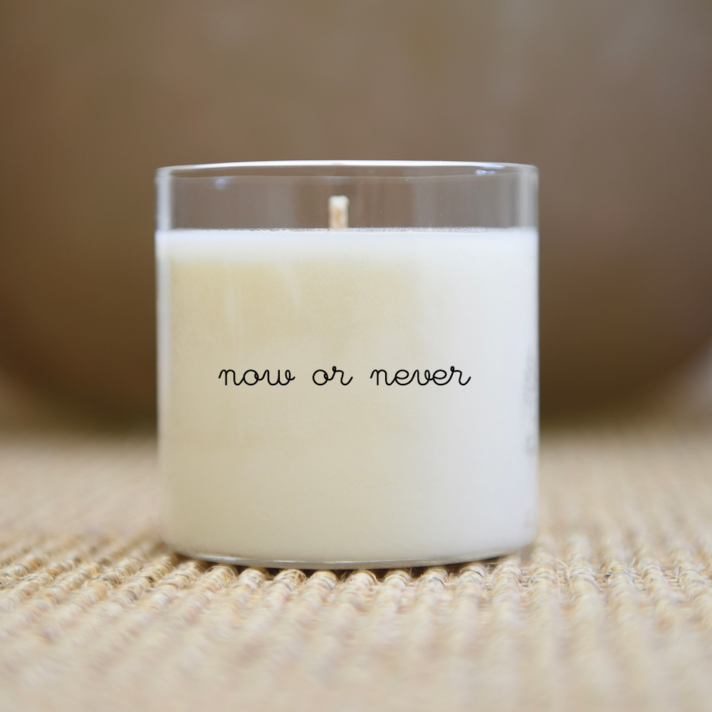 Bougie blanche dans un verre transparent avec le texte « maintenant ou jamais », posée sur une surface texturée.