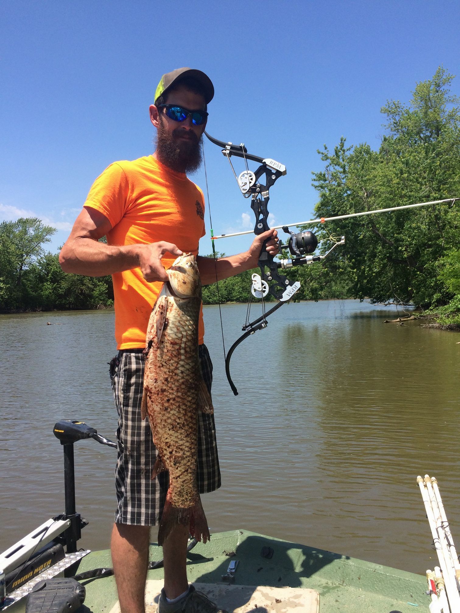 Man in orange shirt holding a large fish, bow in hand, on a boat in a river.