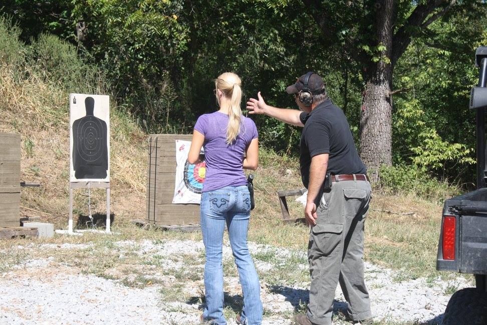 A person in a purple shirt and jeans receives instruction from an instructor at an outdoor shooting range.