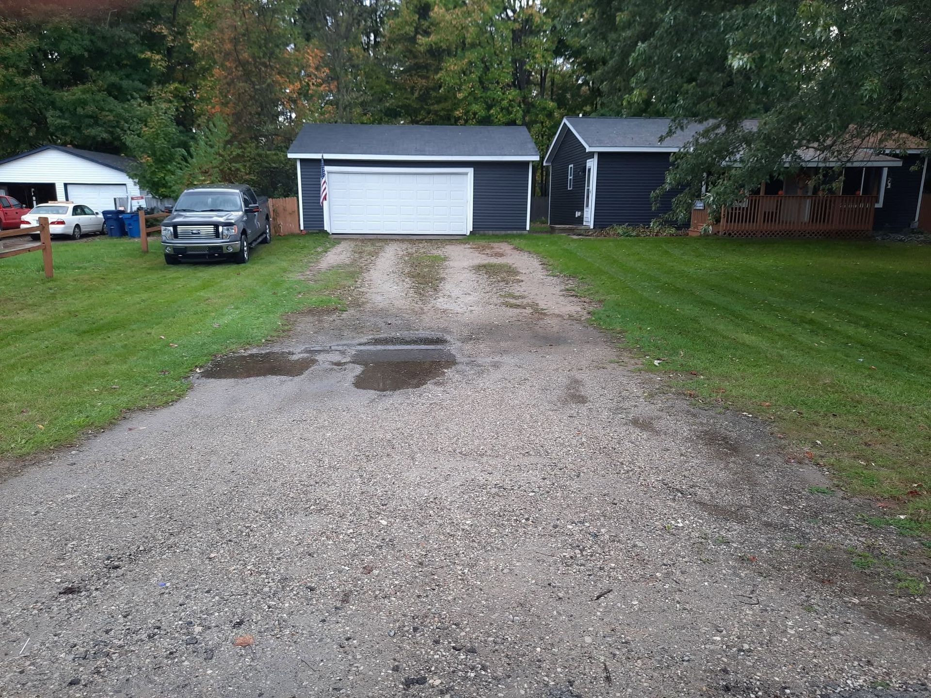 Gravel driveway leading to a gray garage, with a parked car, lawn, trees, and a puddle in the foreground.