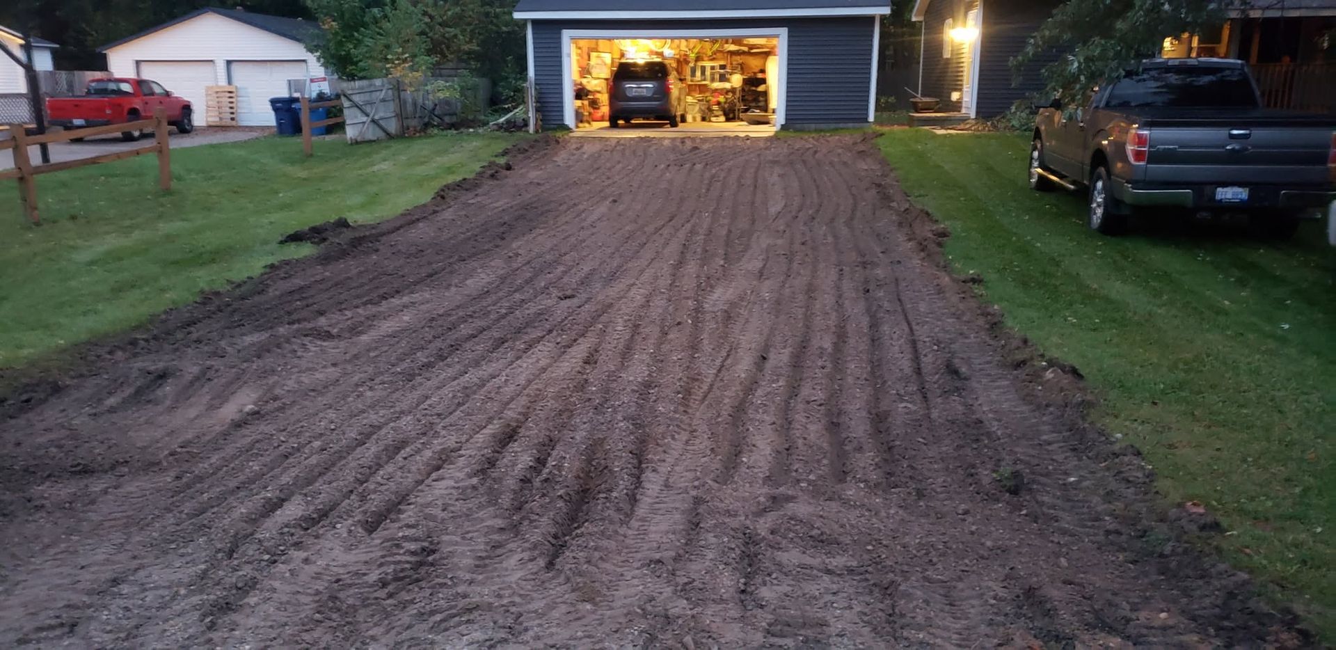 Freshly graded dirt driveway leading to a lit garage beside a parked pickup at dusk