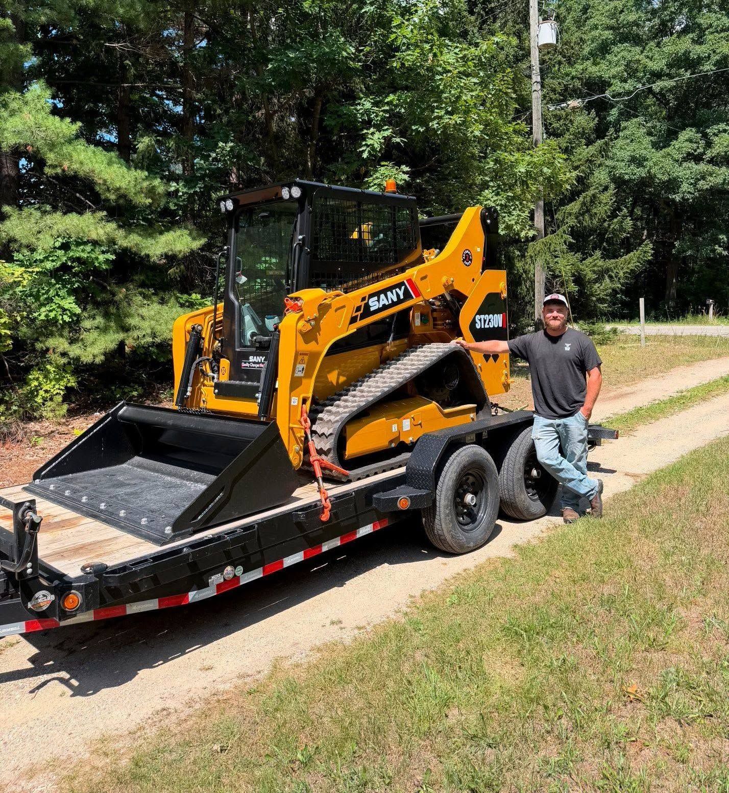 Orange skid steer on a trailer with a man standing beside it on a grassy roadside.