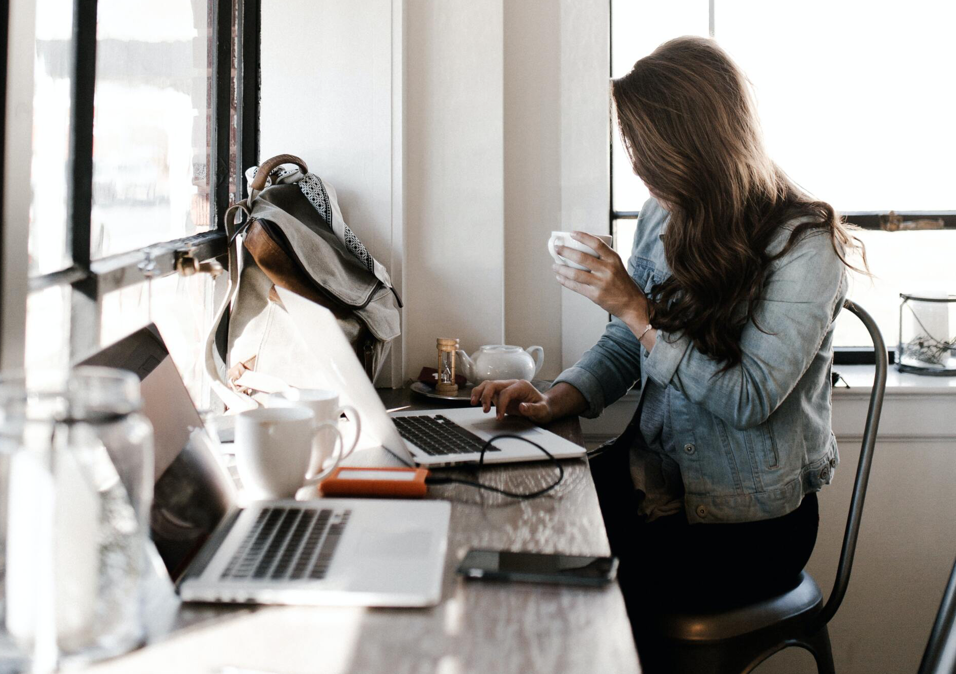 Woman in a denim jacket working on a laptop at a table, holding a cup, and looking at the screen.