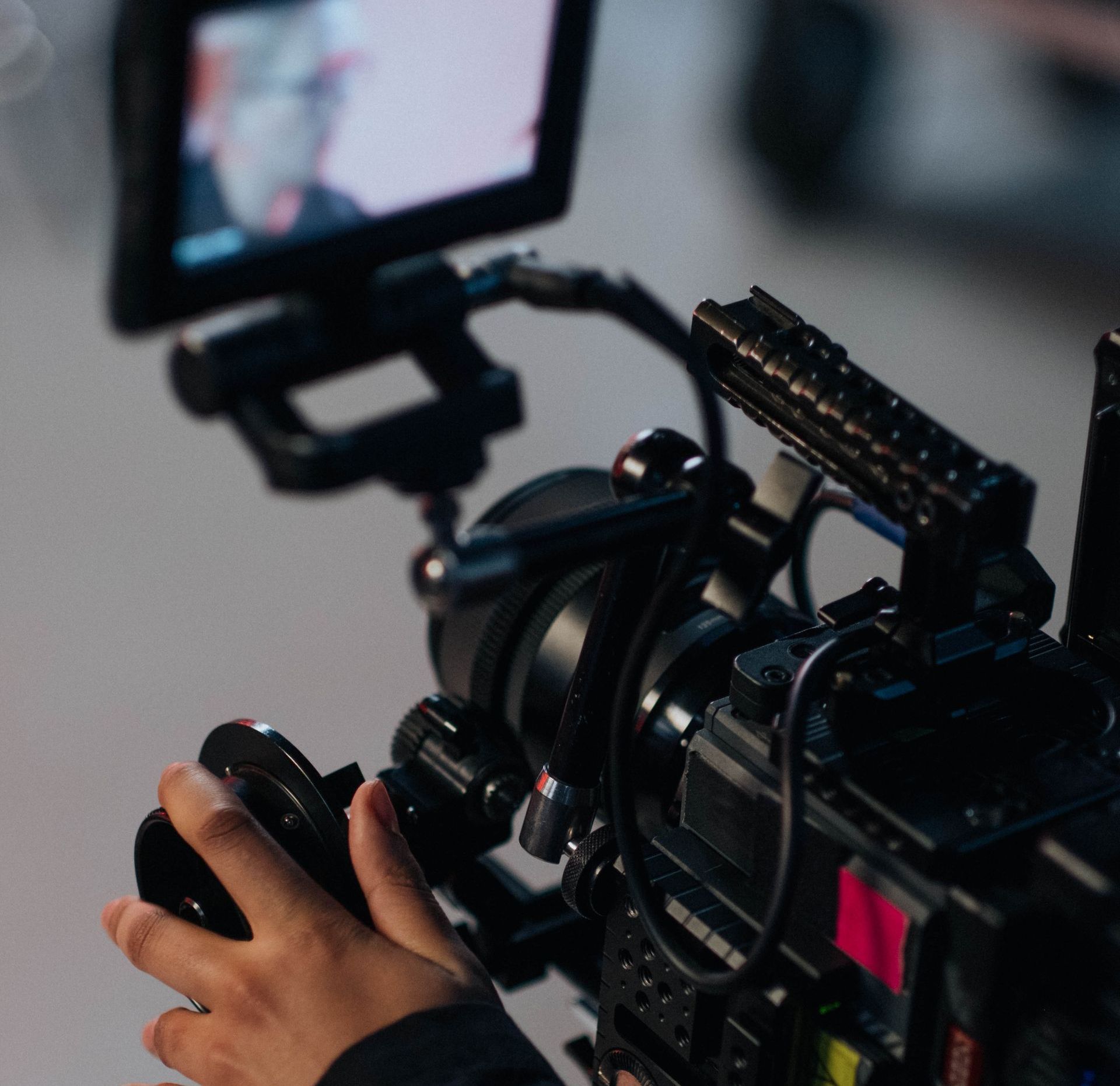 Close-up of a person's hand holding a professional video camera, with an attached screen displaying a blurred image.