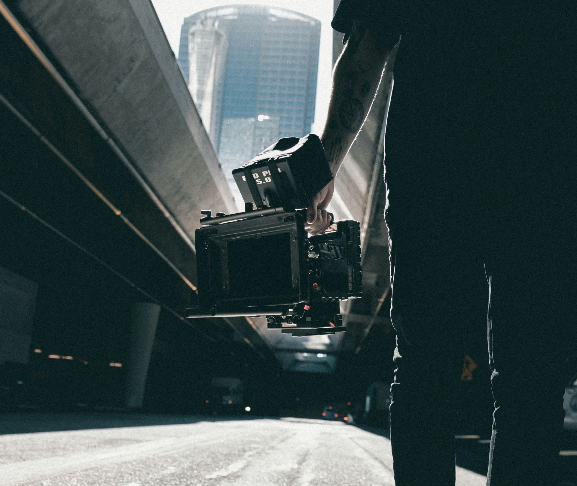 Man carrying a red cinema camera in a city A person is holding a camera in their hand under a bridge