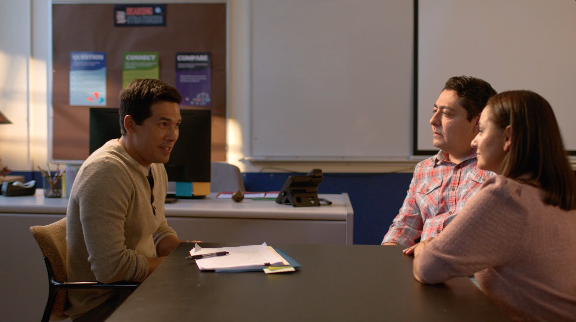 A man in a beige sweater speaks to a couple seated at a table. They are in an office setting.