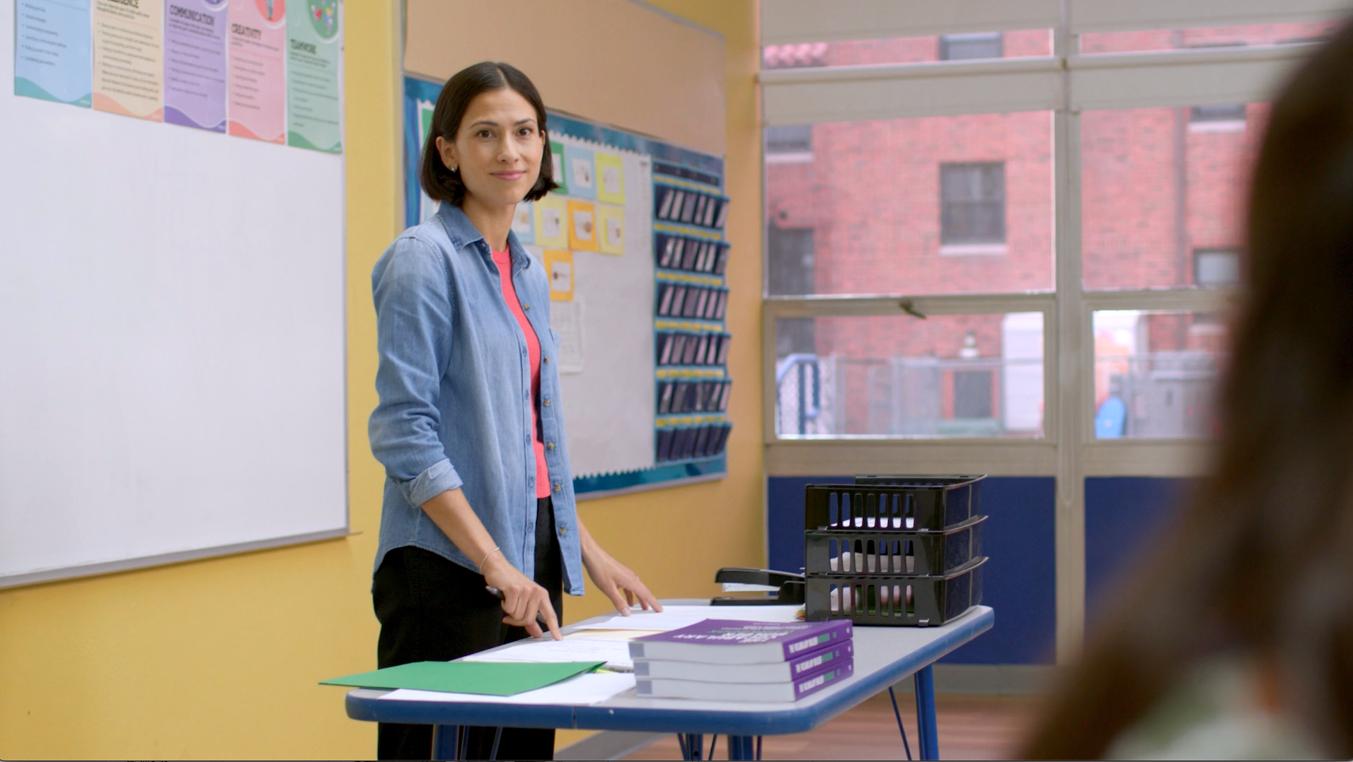 Teacher in a classroom stands behind a desk, smiling. Whiteboard and bulletin board in the background.