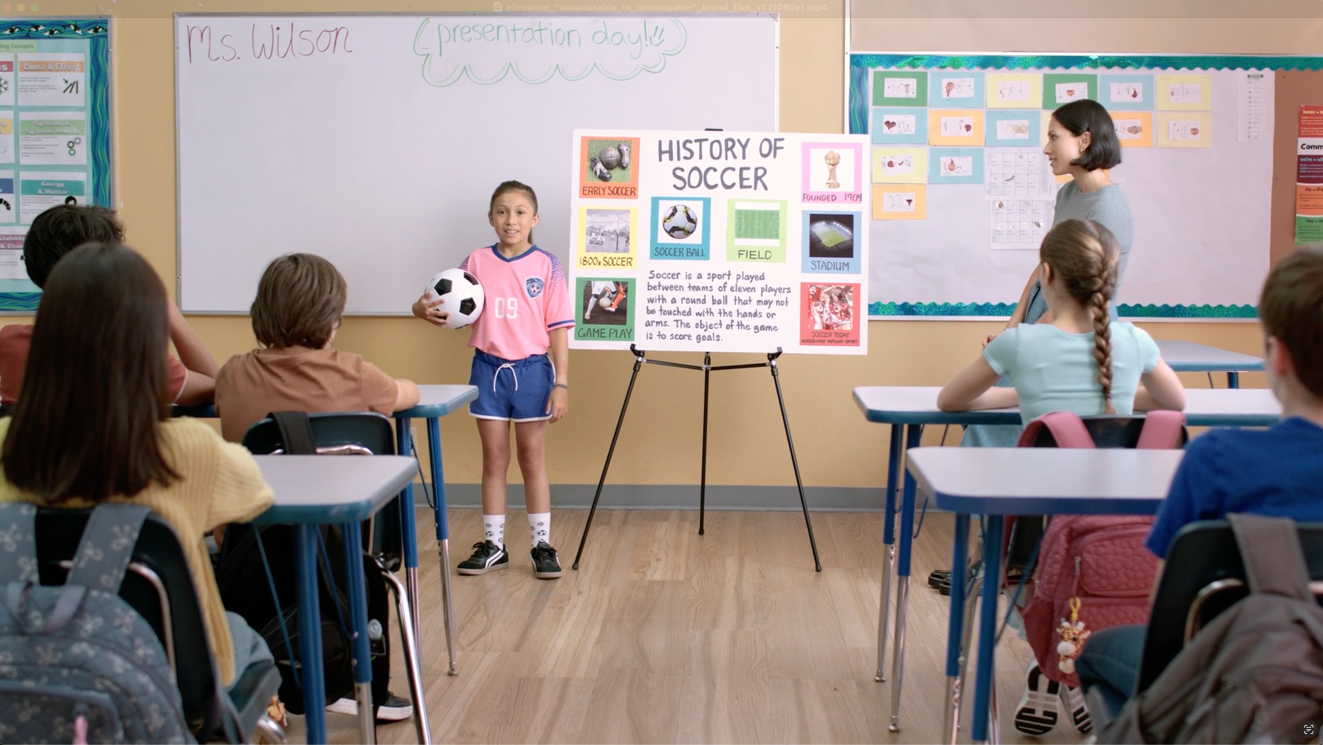 Girl presents soccer report in a classroom. Students seated at desks, teacher stands nearby. Poster visible.