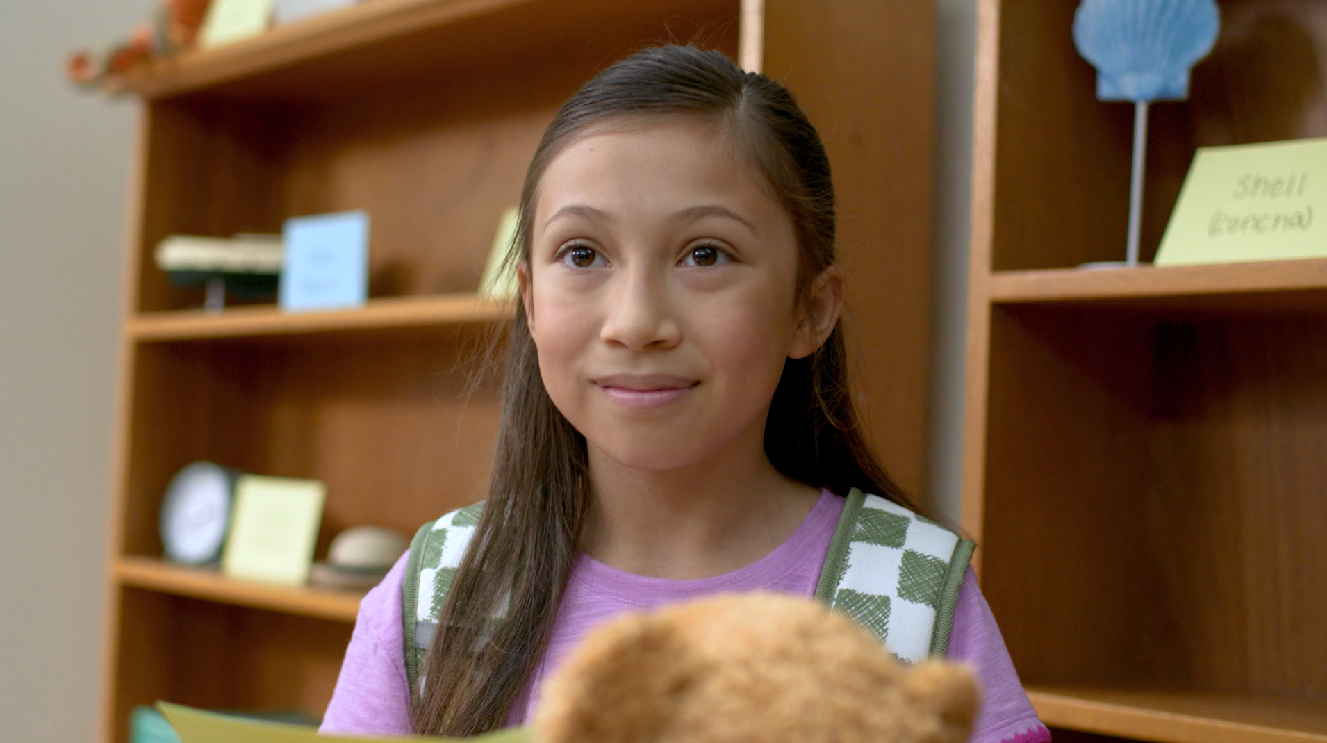 Girl with long brown hair smiles, standing in a classroom. Wooden shelves and a teddy bear in view.