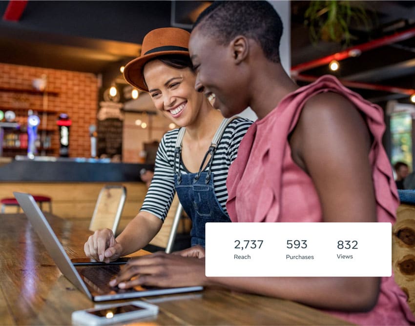 Facebook advertising reach purchases views two women in coffee shop using laptop Two women smile while looking at a laptop in a cafe. The screen displays stats; one woman points.
