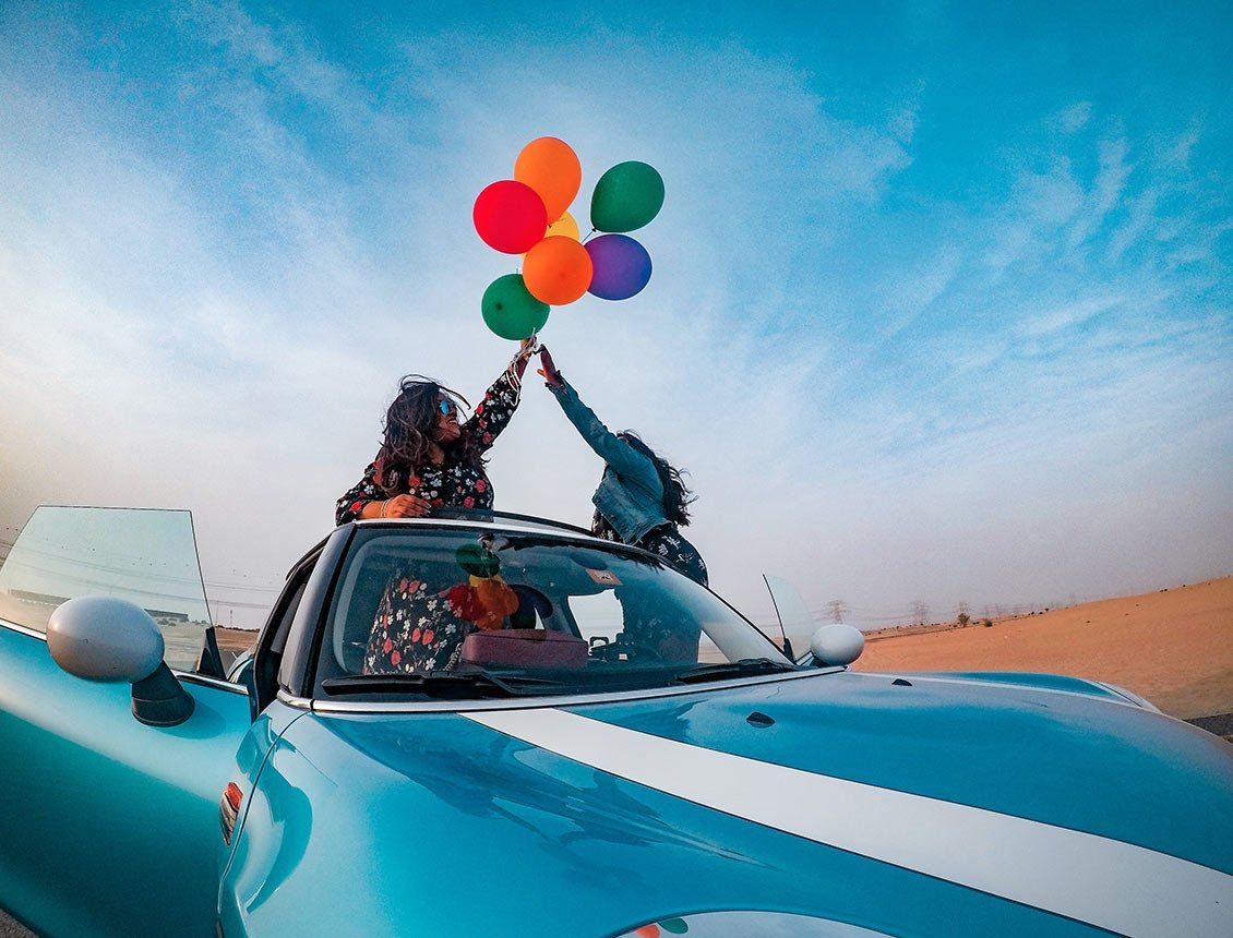 low angle wide shot of two people standing up on convertible car holding baloons A woman is holding balloons while sitting in the back of a car.