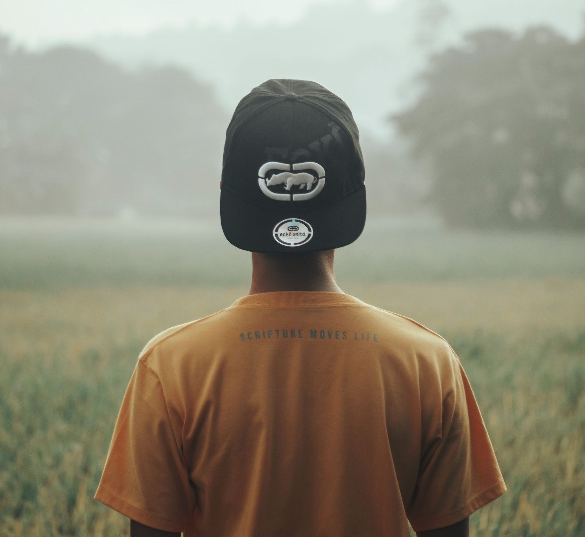 Image of a person from behind wearing hat backwards looking out onto a foggy field A man wearing a hat with the letter g on it