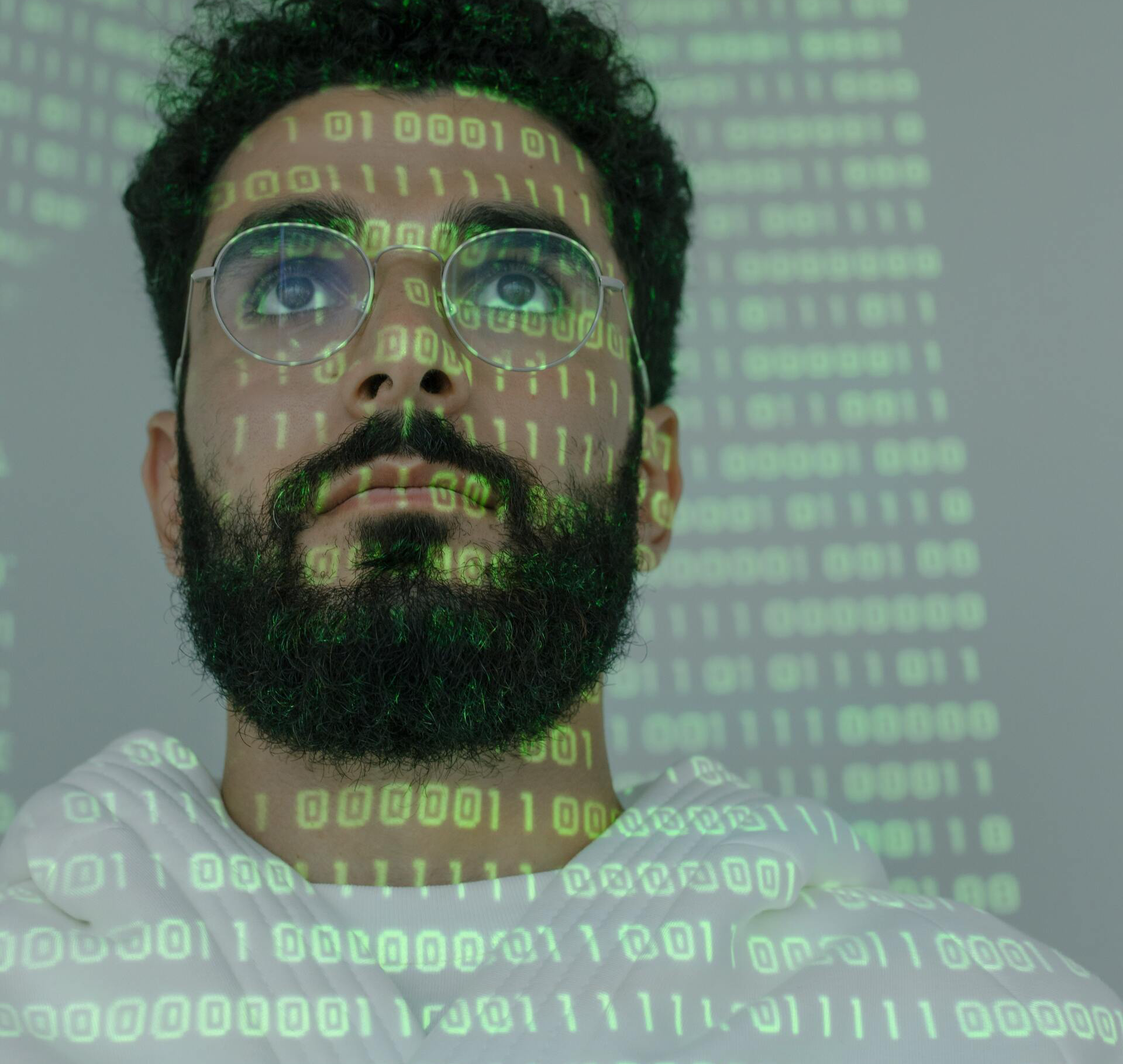 Close up man looking up wearing glasses with ones and zeroes projected on his face and clothing A man with glasses and a beard is surrounded by binary code