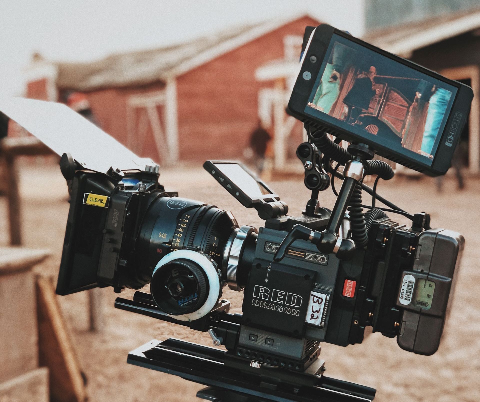 RED Dragon cinema camera old western town location A professional video camera on a tripod, in front of a red building and blurred background.