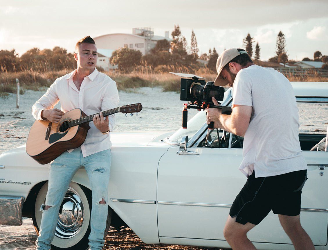 behind the scenes image of a videographer filming a man playing a guitar in front of car behind the scenes image of a videographer filming a man playing a guitar in front of car