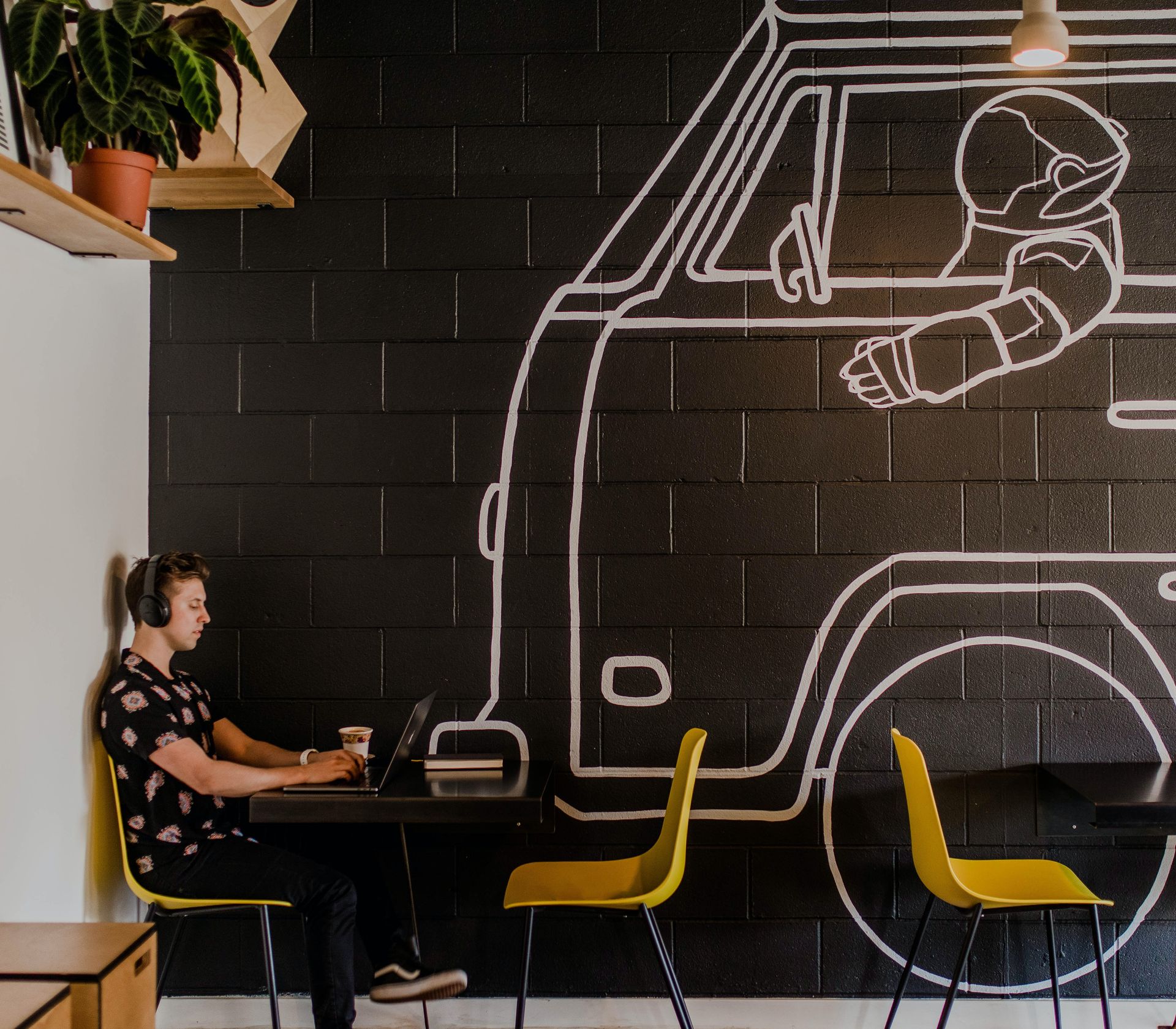 Man working on laptop in creative environment Man using a laptop at a table in a cafe with a van mural and yellow chairs.