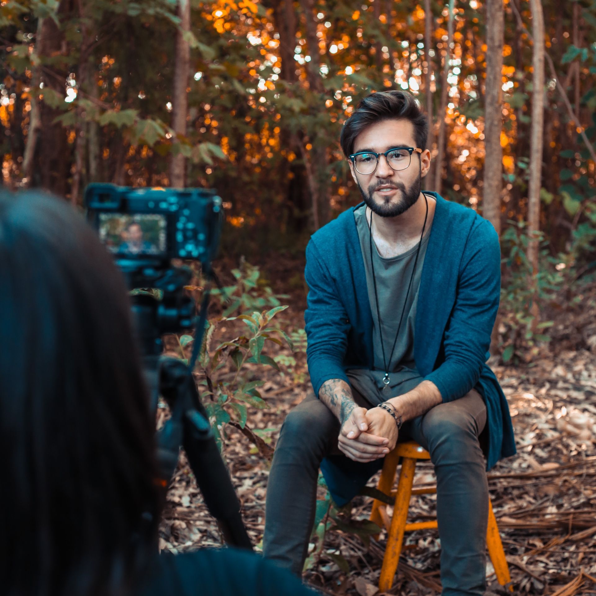 Behind the scenes testimonial video shoot Man sitting in front of a camera in a forest setting, being filmed. He wears glasses and a blue cardigan.