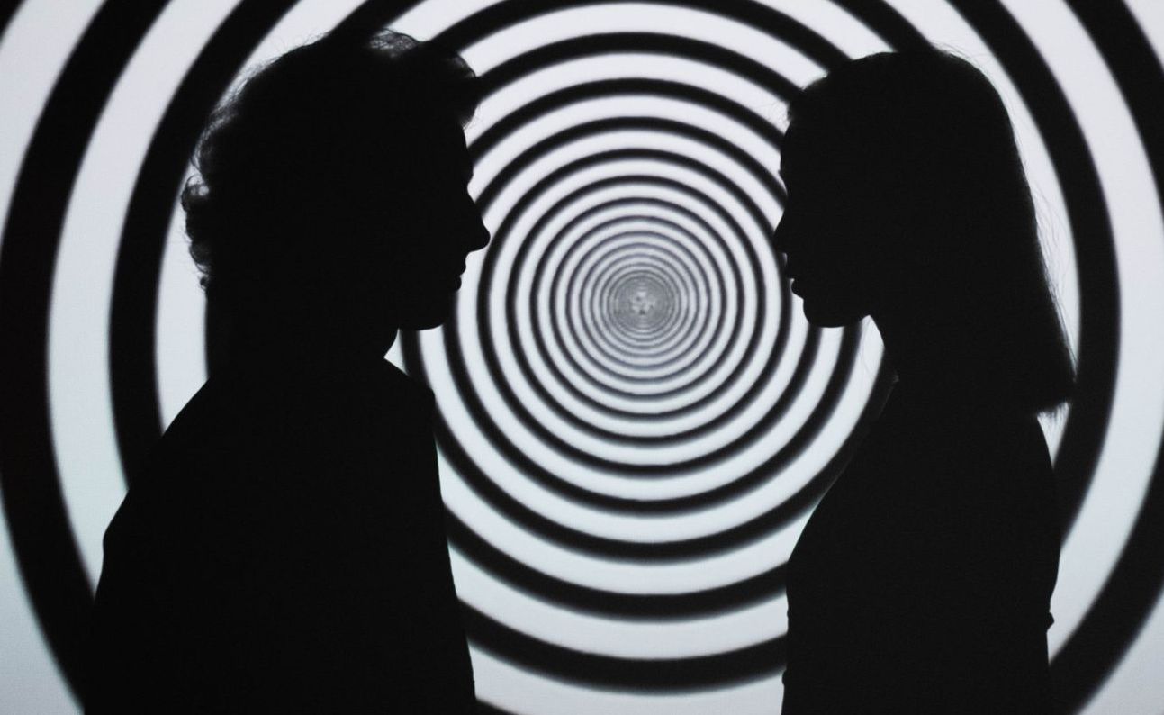 Couple facing each other in silhouette with rings of black and white behind them Silhouetted figures face each other before a swirling black and white spiral.