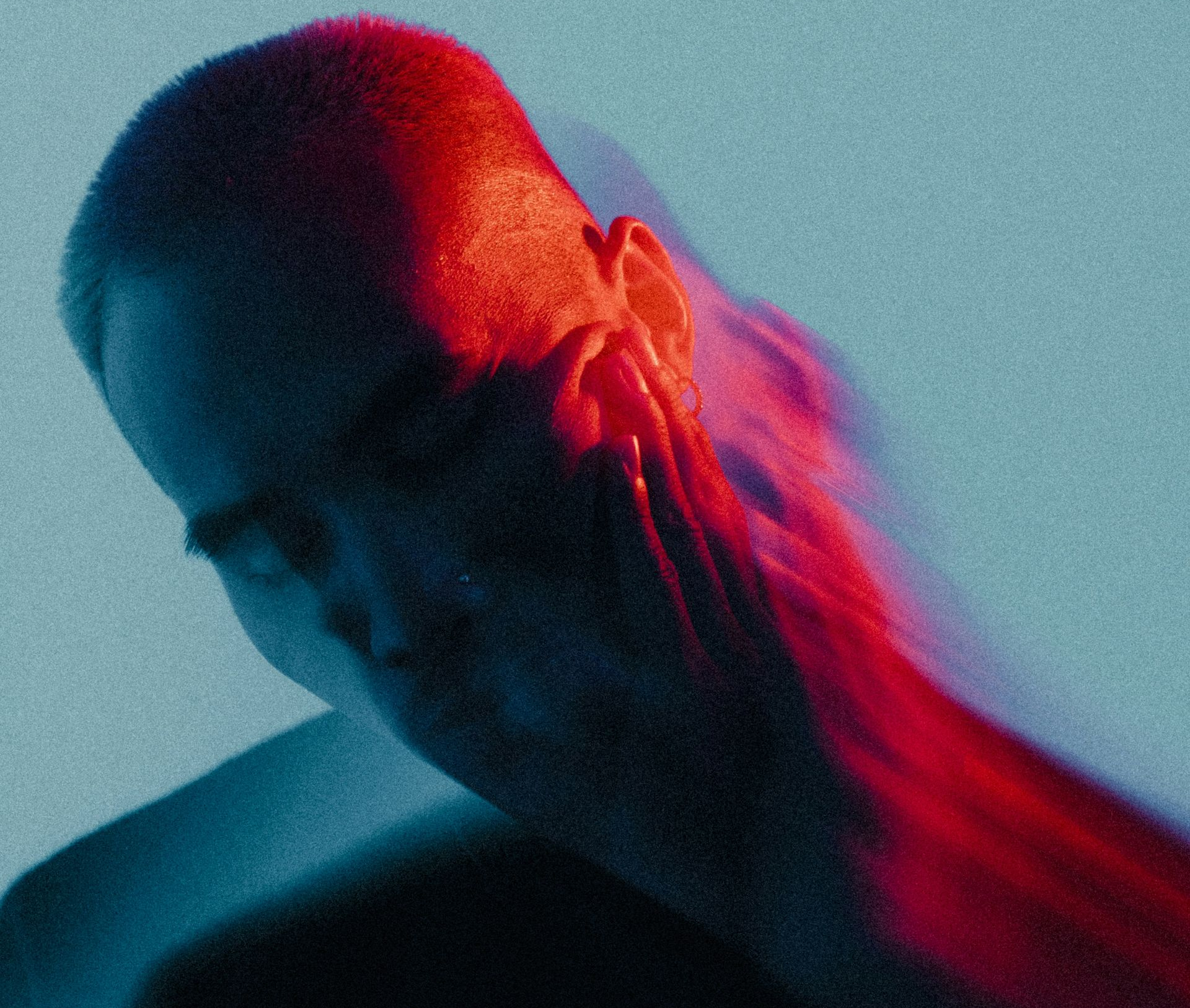 A person's head and shoulder illuminated by red and blue light, with a blurred silhouette. Person with close-cropped hair, lit with red and blue light, head and shoulders visible, looking down.