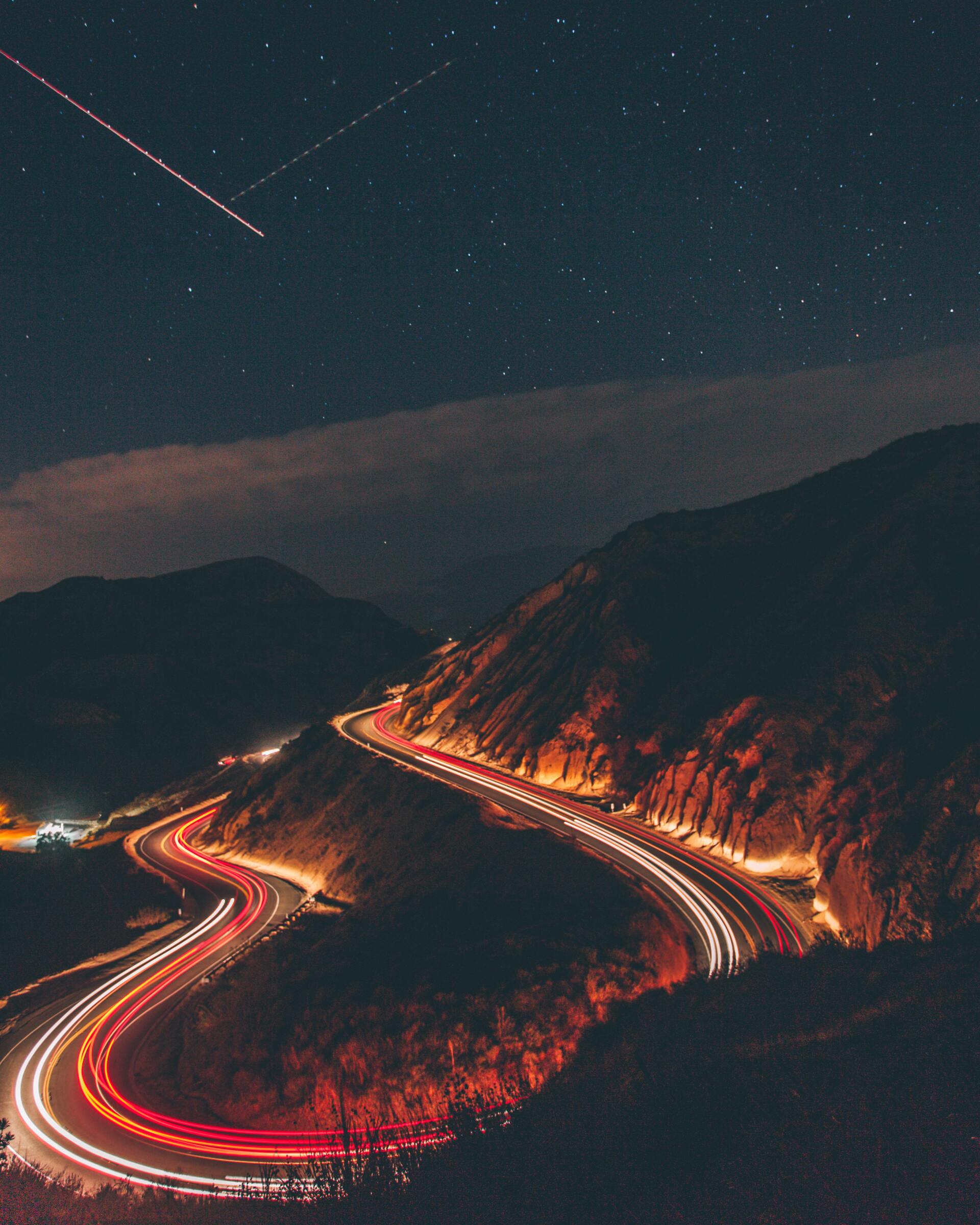 image of time lapse cars driving on mountain with shooting stars in sky Winding road with streaks of car lights through dark mountains under a starry night sky with a shooting star.