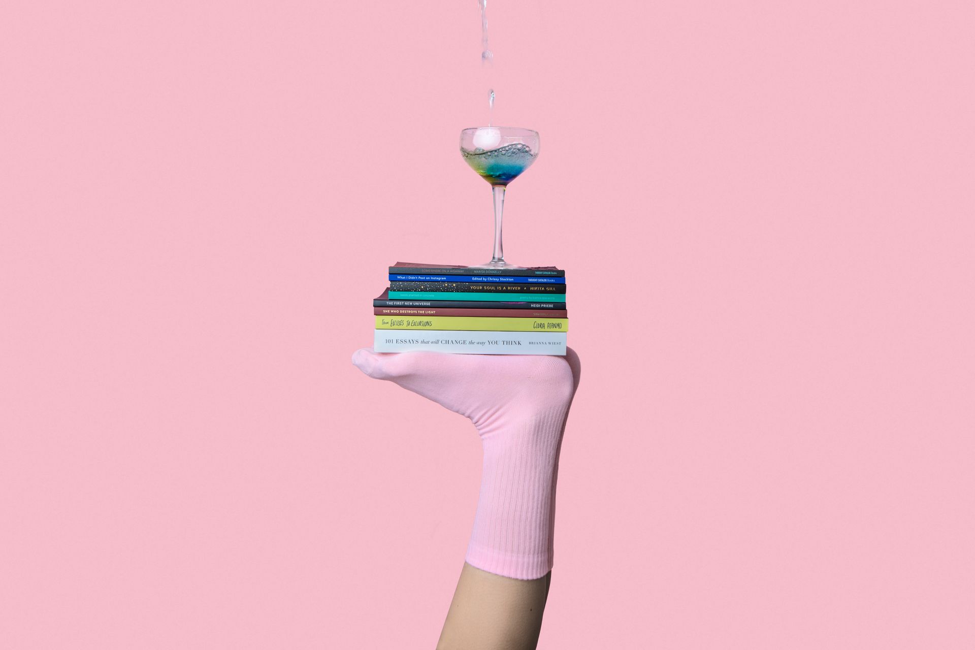 A hand in a pink sock is holding a stack of books and a glass of wine.