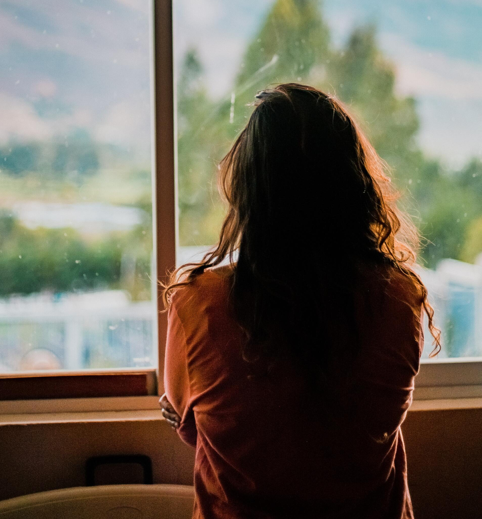 A woman in a red shirt is looking out a window
