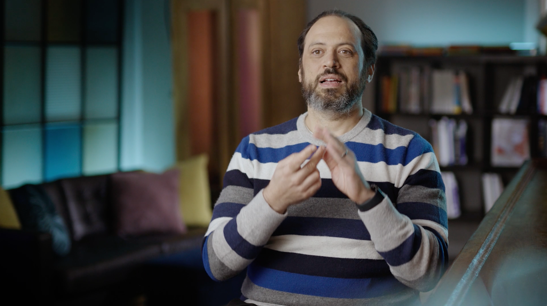 Man with a beard in striped sweater gestures with hands; interior with couch and bookshelf.
