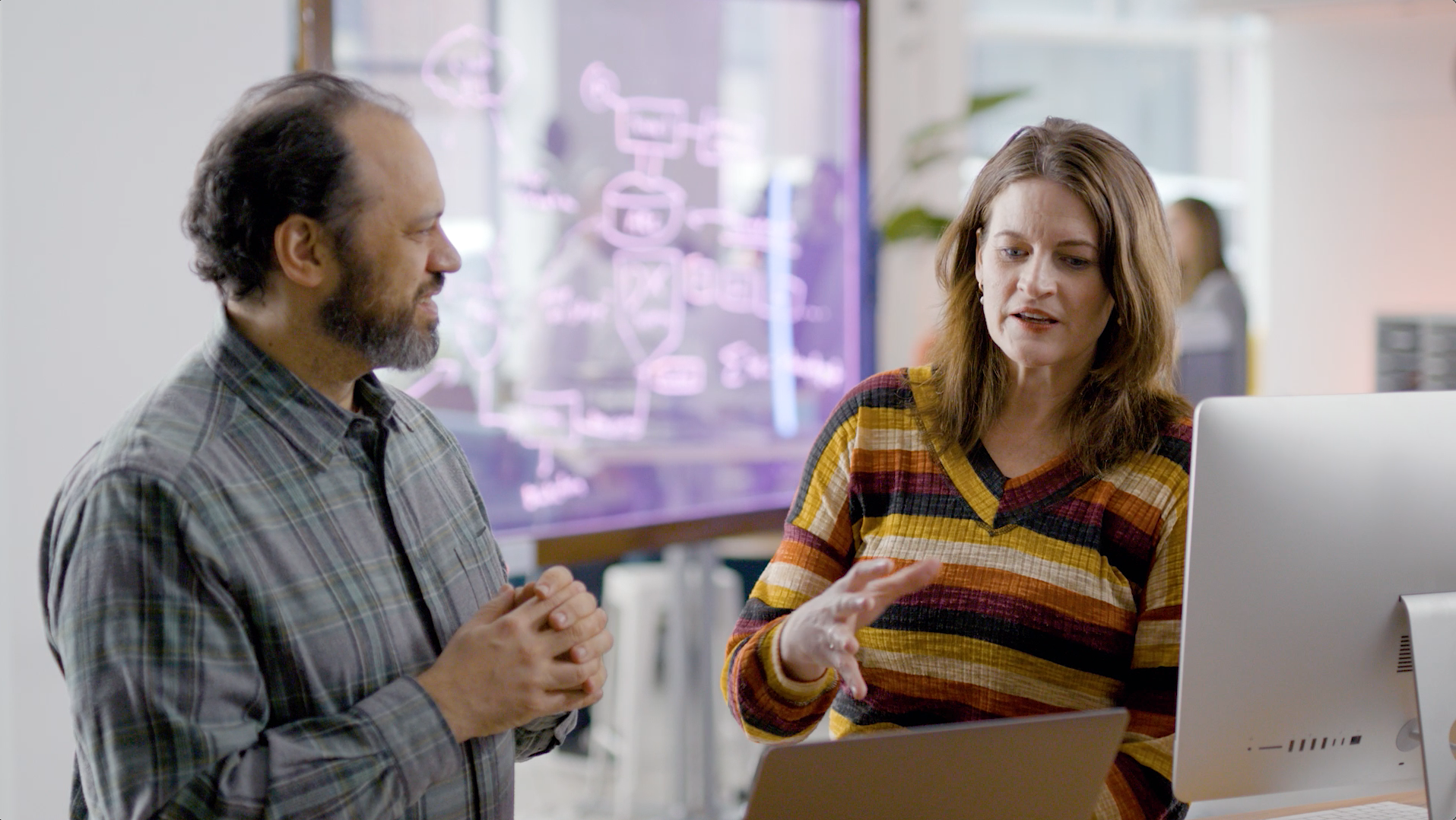 Two colleagues discussing a project at a computer, one gesturing with hand.