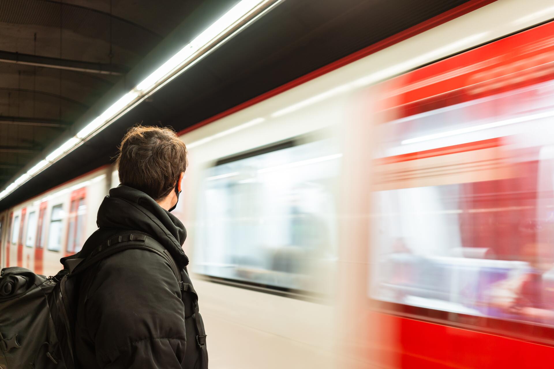 Image from behind of man wearing a mask at subway platform as a subway goes by in motion blur A man is waiting for a train at a subway station.