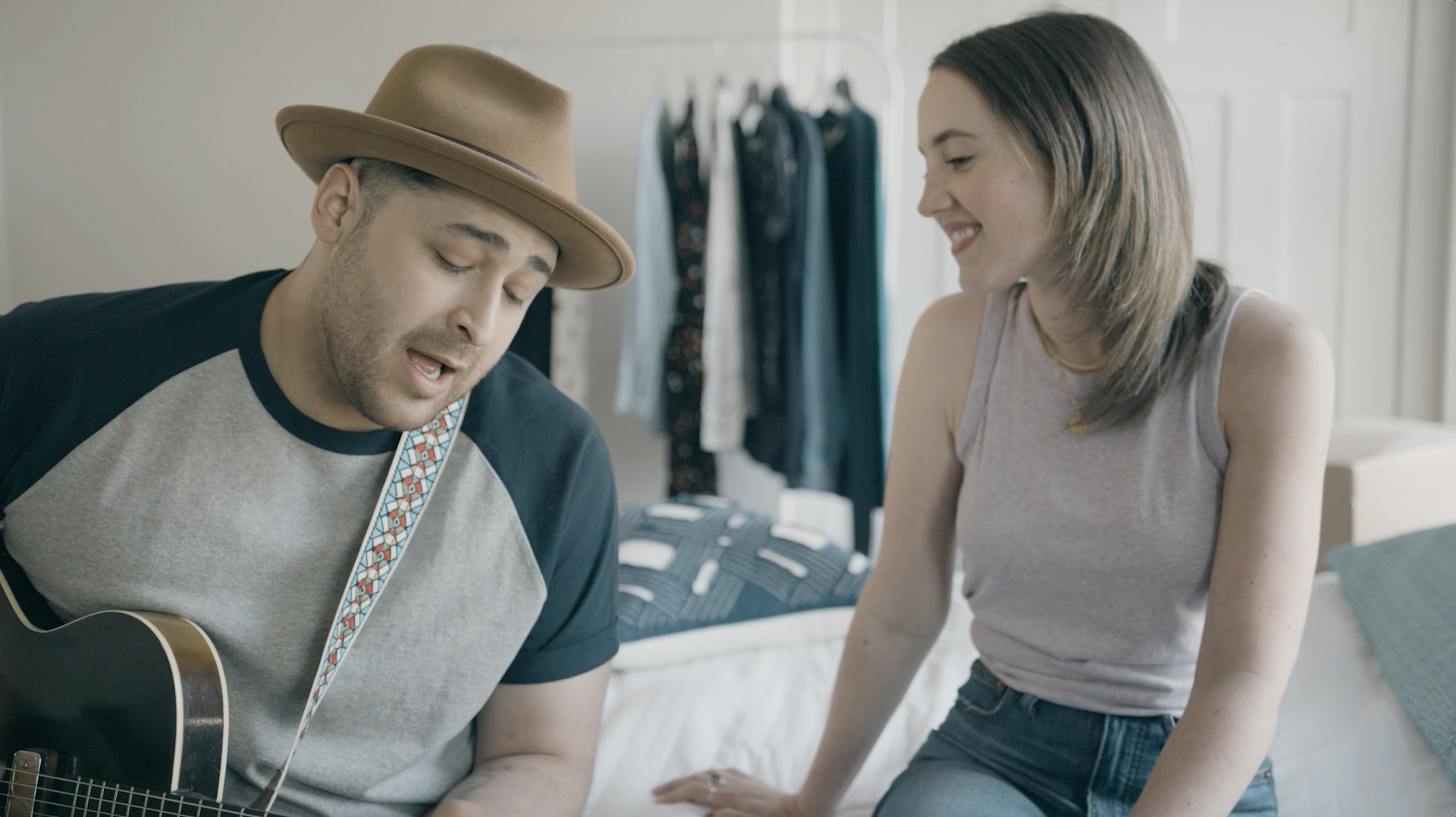 A man is playing a guitar for a woman while they are sitting on a bed.