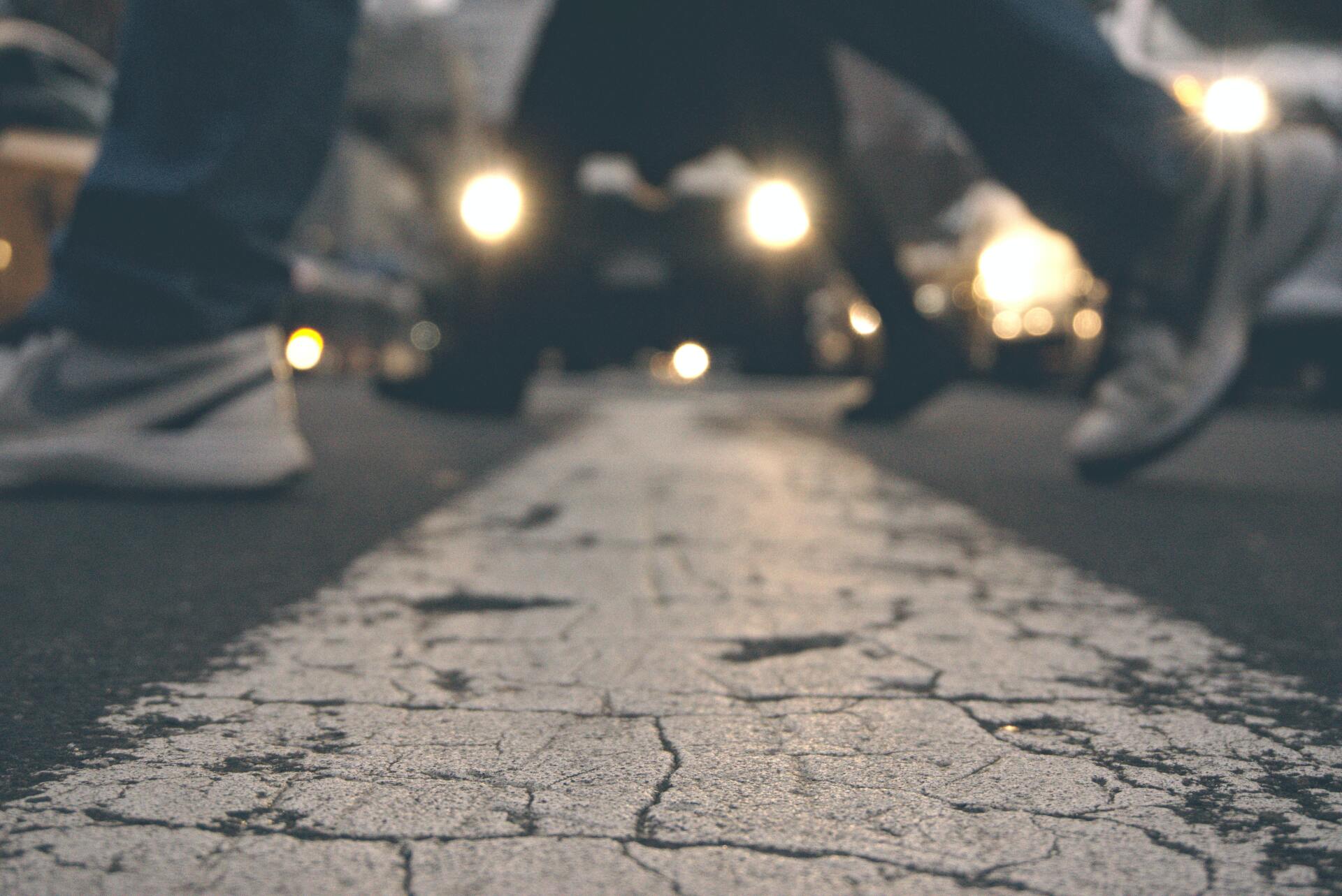 close up people's feet crossing street at dusk People walking on a crosswalk in a city at night, car headlights blurred in the background.