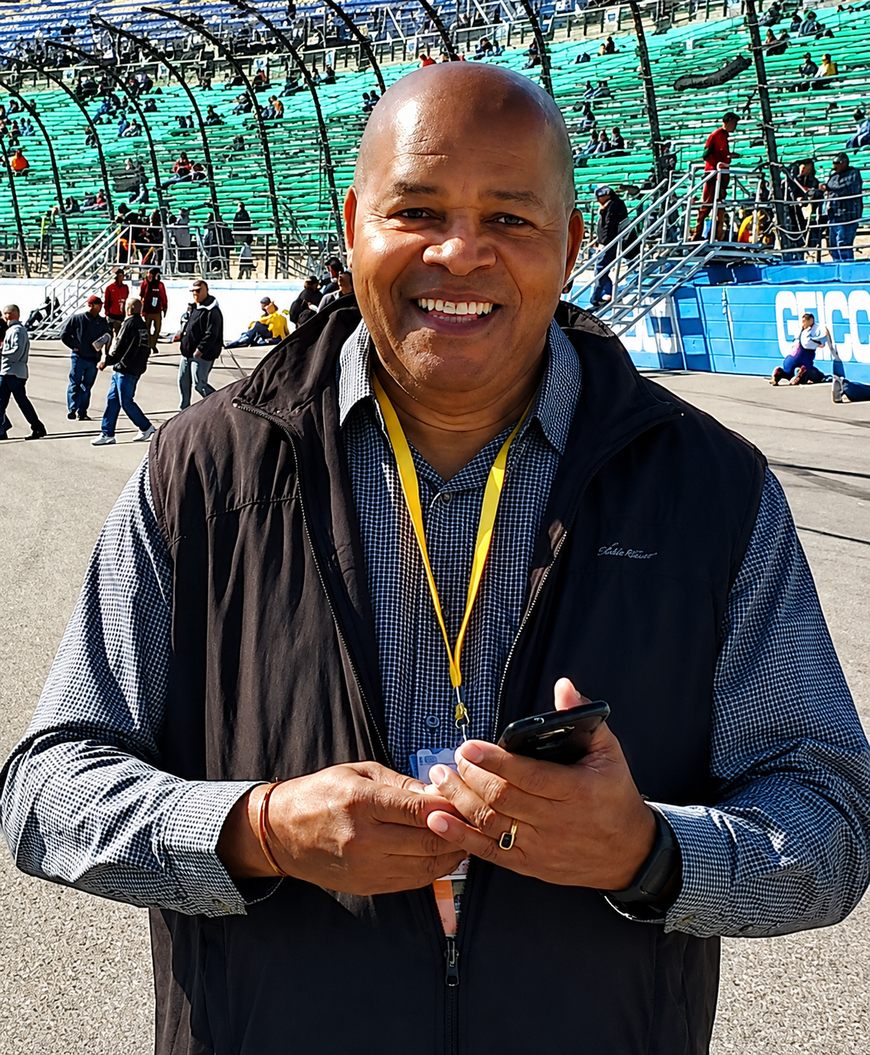 Smiling person wearing a lanyard, holding a phone, standing in a crowded outdoor stadium.