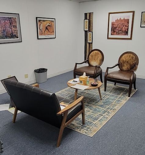 Small office seating area with black sofa, two wooden chairs, coffee table, rug, and framed art on white walls