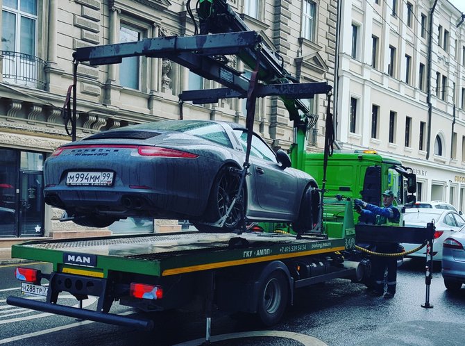 Car on a tow truck in a city street, with a green crane lifting it amid parked vehicles.
