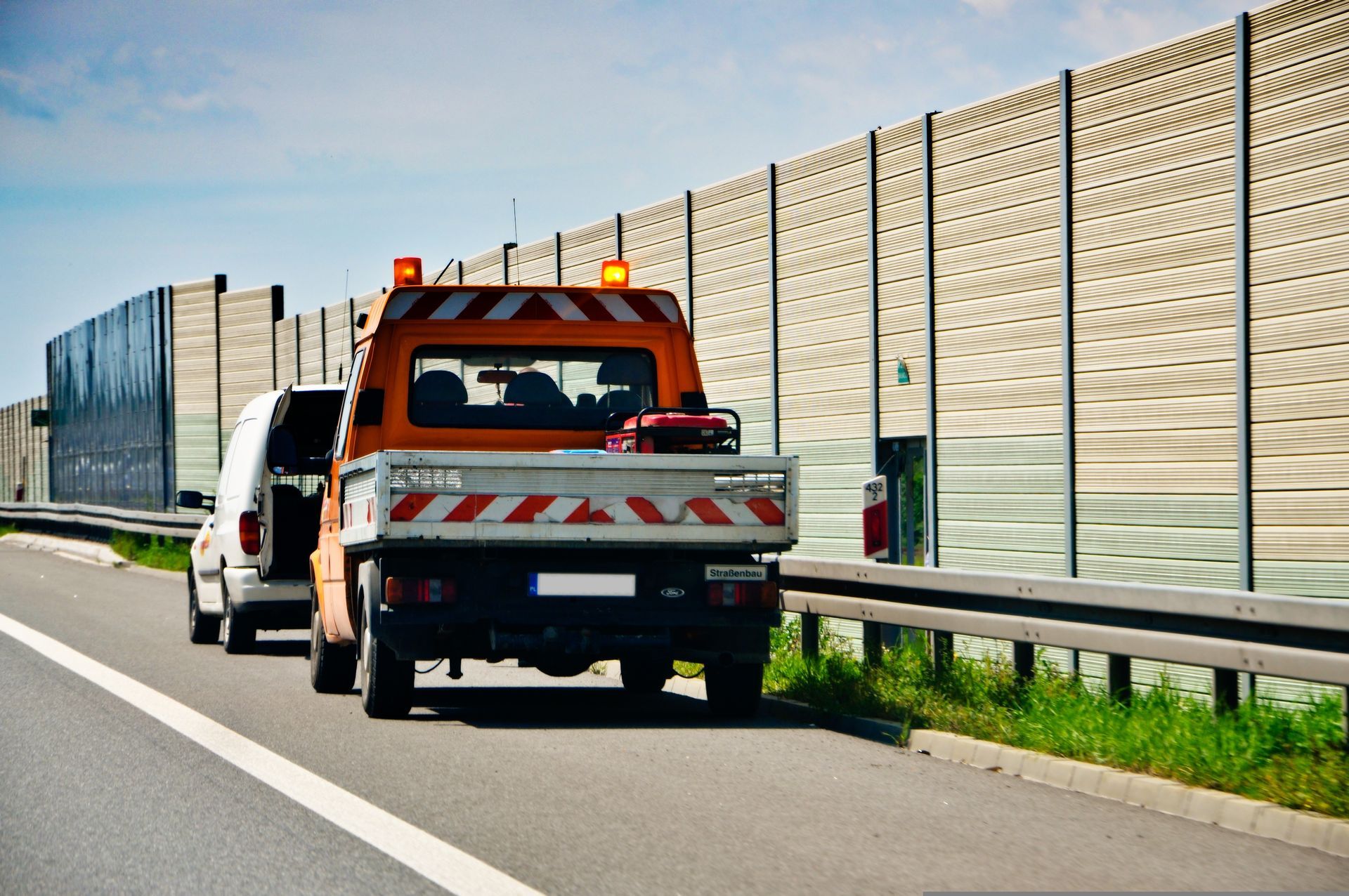 Orange tow truck on roadside beside tall sound barrier and guardrail on a highway