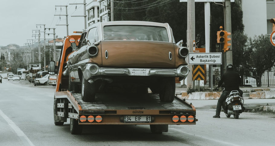 Tow truck hauling a vintage brown car on a city street, with traffic and road signs in the background