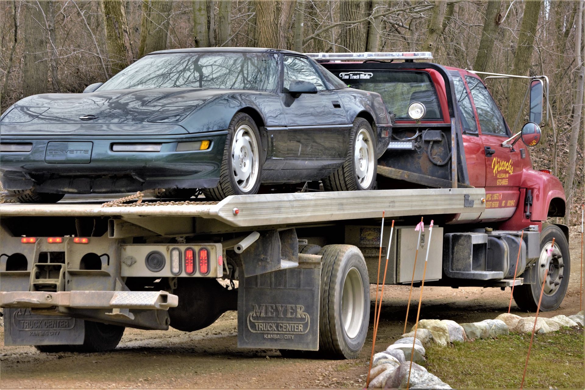 Sports cars loaded on a flatbed tow truck beside a red semi-truck on a roadside.