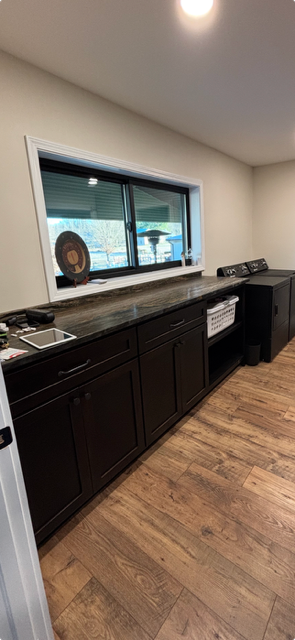 Empty kitchen with dark cabinets, wood floor, sink, window, and coffee maker under bright lighting