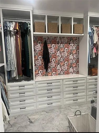 Built-in walk-in closet with white drawers, hanging clothes, shelves, and floral wallpaper back panel