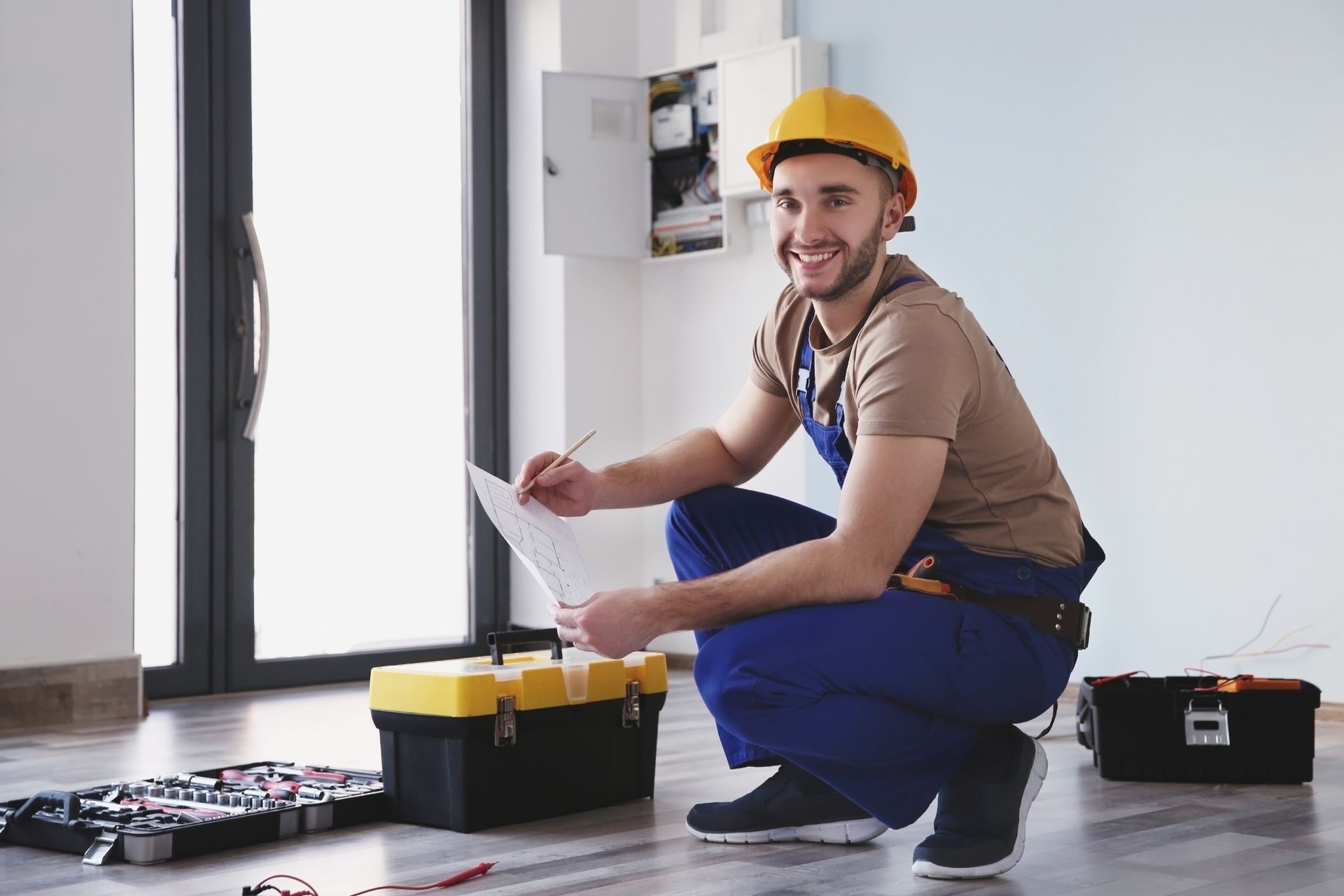 Worker in yellow hard hat kneeling with tools in a bright room, checking electrical wiring. Worker in yellow hard hat kneeling with tools in a bright room, checking electrical wiring.