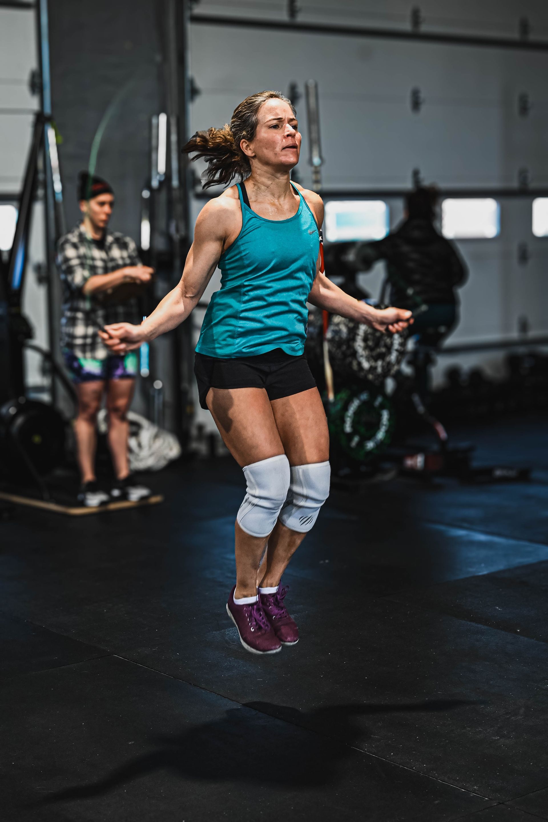 Woman in gym jumps rope, wearing knee pads, black shorts, turquoise top. Another person in background.